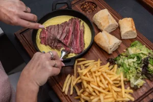 Hands slicing tender steak in signature entrecôte sauce with fries, salad and warm baguette at Memories L’Entrecôte’s Restaurant Rooftop, Rise Urban Art Hotel, Larnaca Cyprus